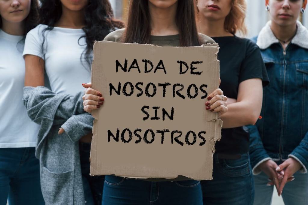 grupo de personas de pie, la mujer del centro sostiene un cartel en un trozo de cartón que dice NADA DE NOSOTROS SIN NOSOTROS